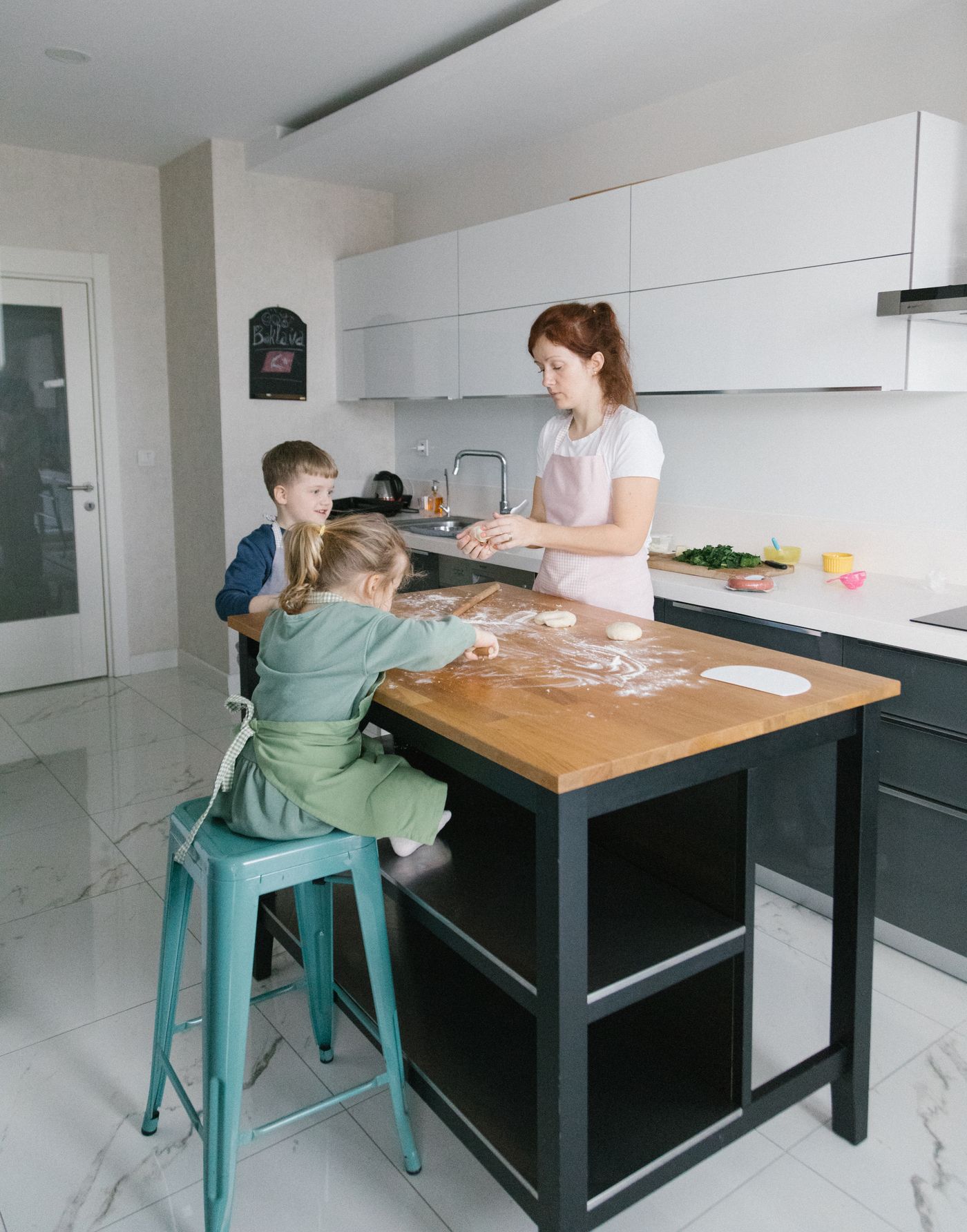 A woman and two small children baking together at a kitchen island dusted with flour