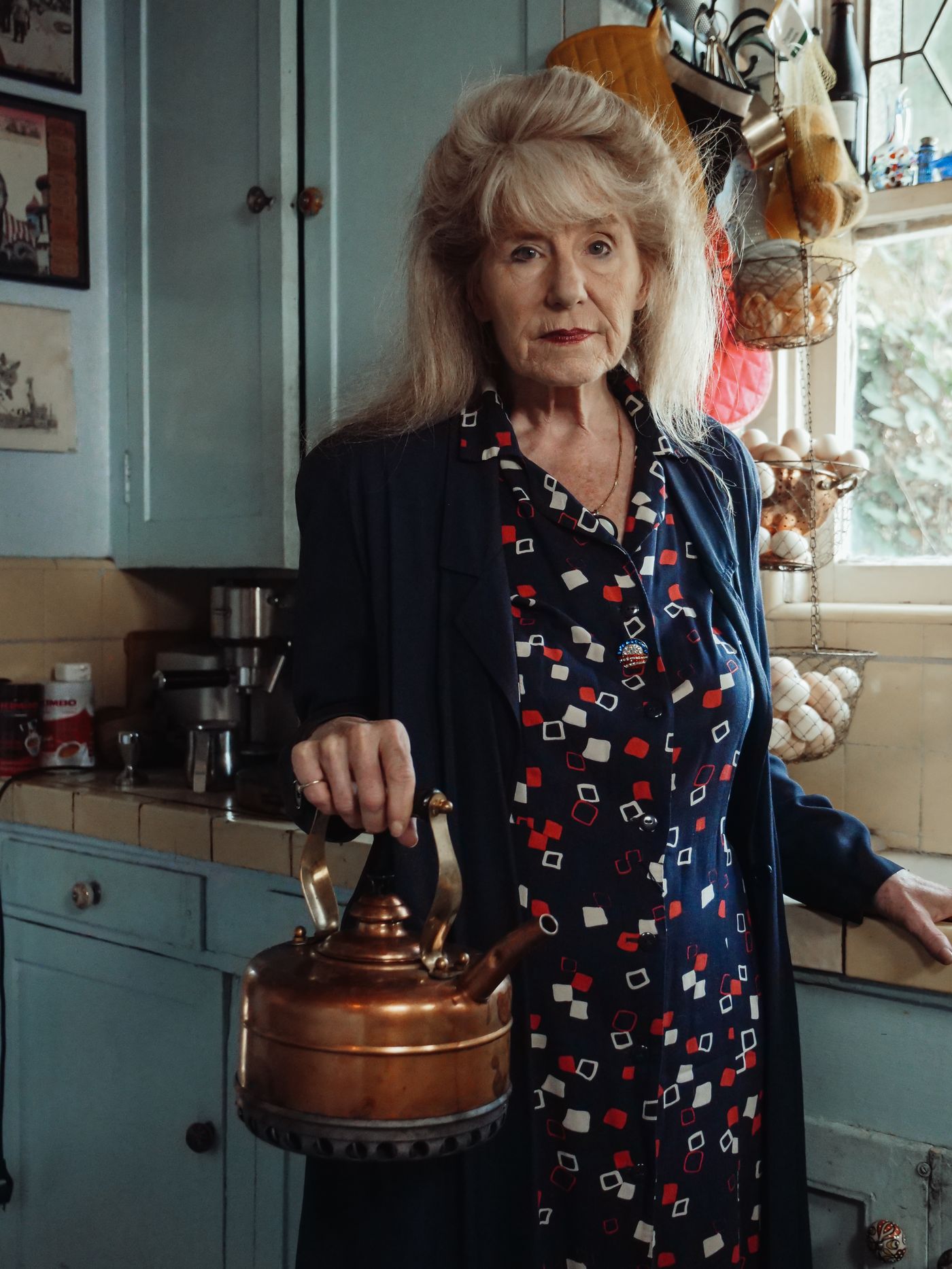 An older woman standing in a well-used kitchen holding a copper kettle